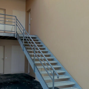 Escalier extérieur en métal galvanisé sans contremarches. Marches tôles métal perforées antidérapantes. Rampe et garde corps : main courante cylindrique, poteaux plats, lisses métalliques droites et soubassement en verre.