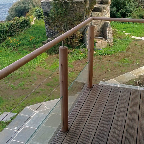 Garde corps d'un escalier en bois Iroko de teinte naturelle d'une maison en bord de mer. Lames striées, poteaux et main courante cylindriques. Pitons et lisses en inox. Soubassement en verre.